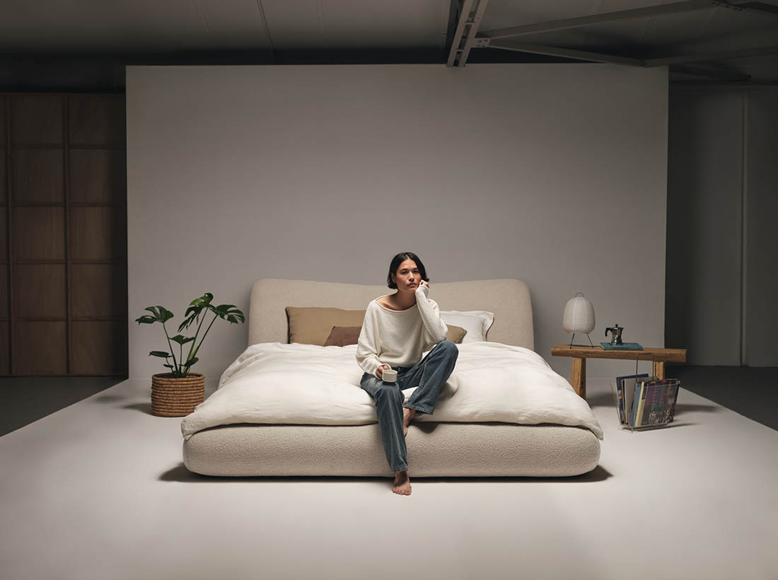 Woman seated on a low beige curved bed in a minimal bedroom with timber side table, plant and soft lighting.