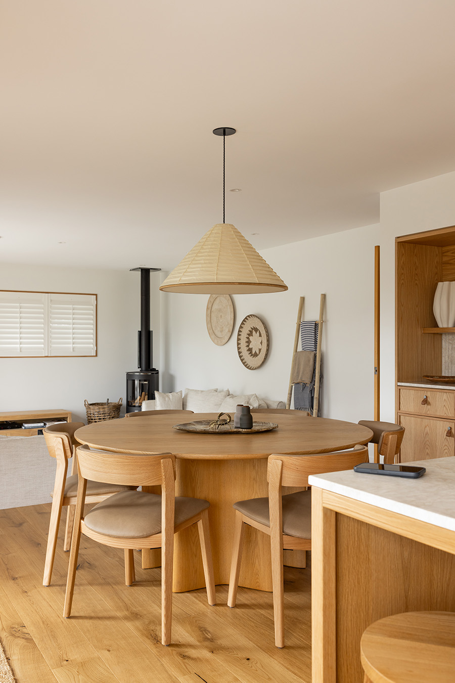 Round oak dining table with sculptural open base styled beneath a woven pendant in an open-plan living and dining room.