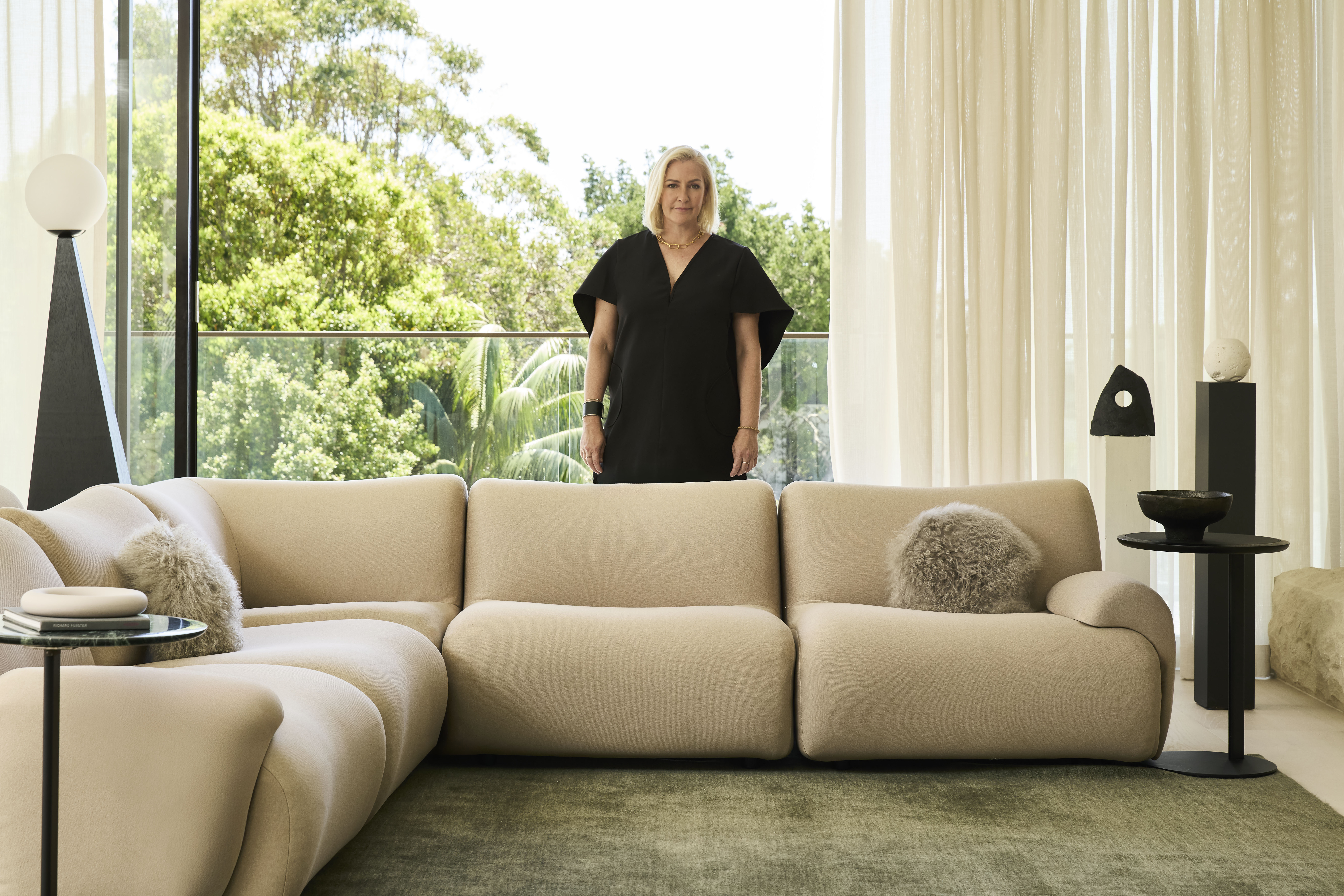 Interior Designer Lara Hutton stands behind a modular sofa in a contemporary living room. The space features neutral upholstery, sculptural decoration, and black accent tables.