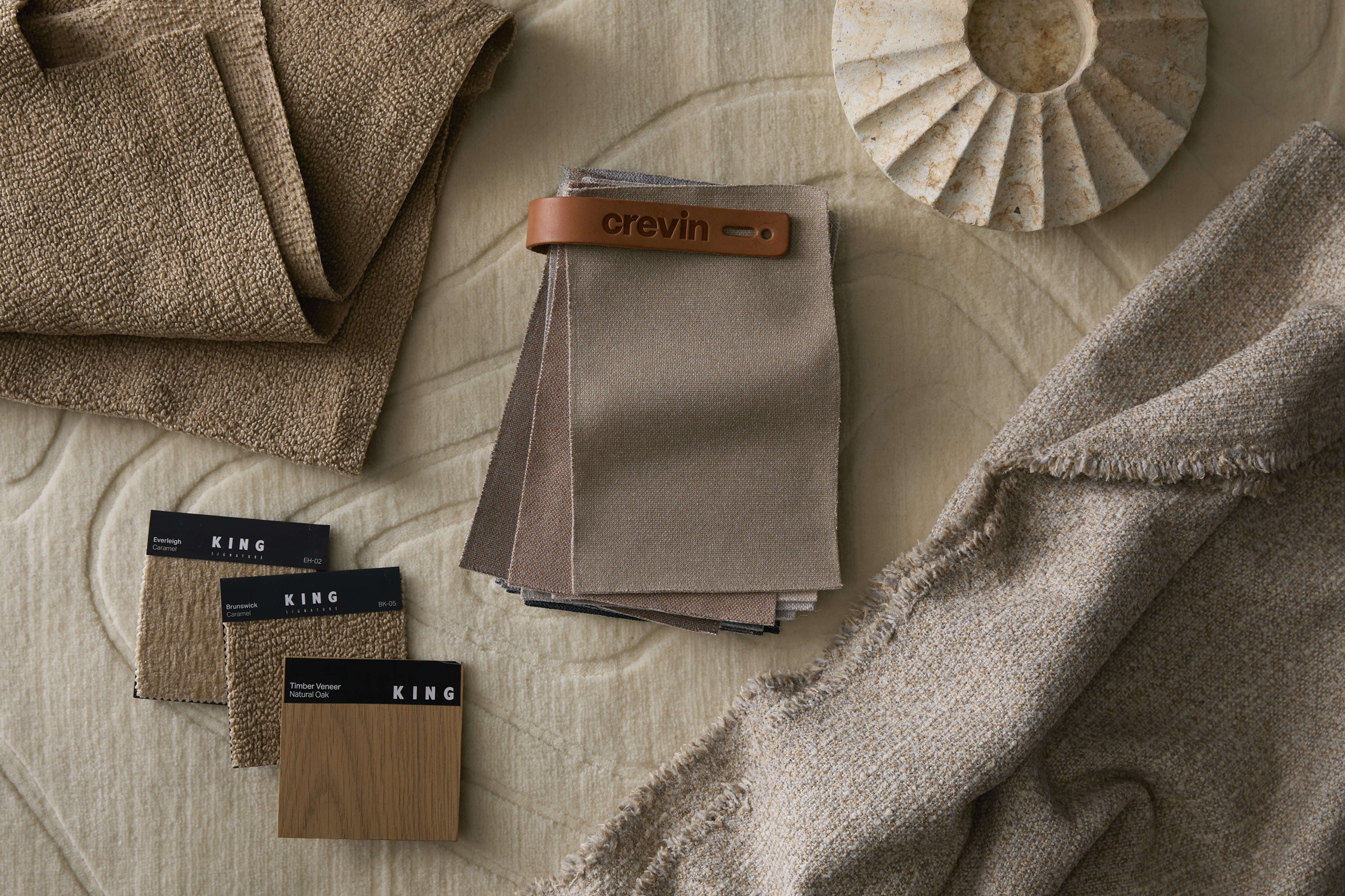 A flat lay of fabric and timber swatches in warm neutral tones, arranged on a textured rug with sculptural decor.