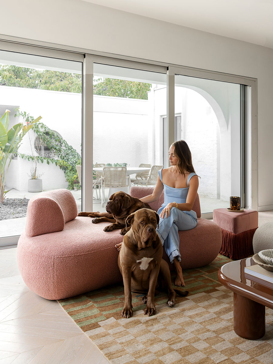 Kate and her dogs sit on a rose-toned sofa in a light-filled living space, complemented by a checkered rug and floor-to-ceiling windows opening to an outdoor courtyard.