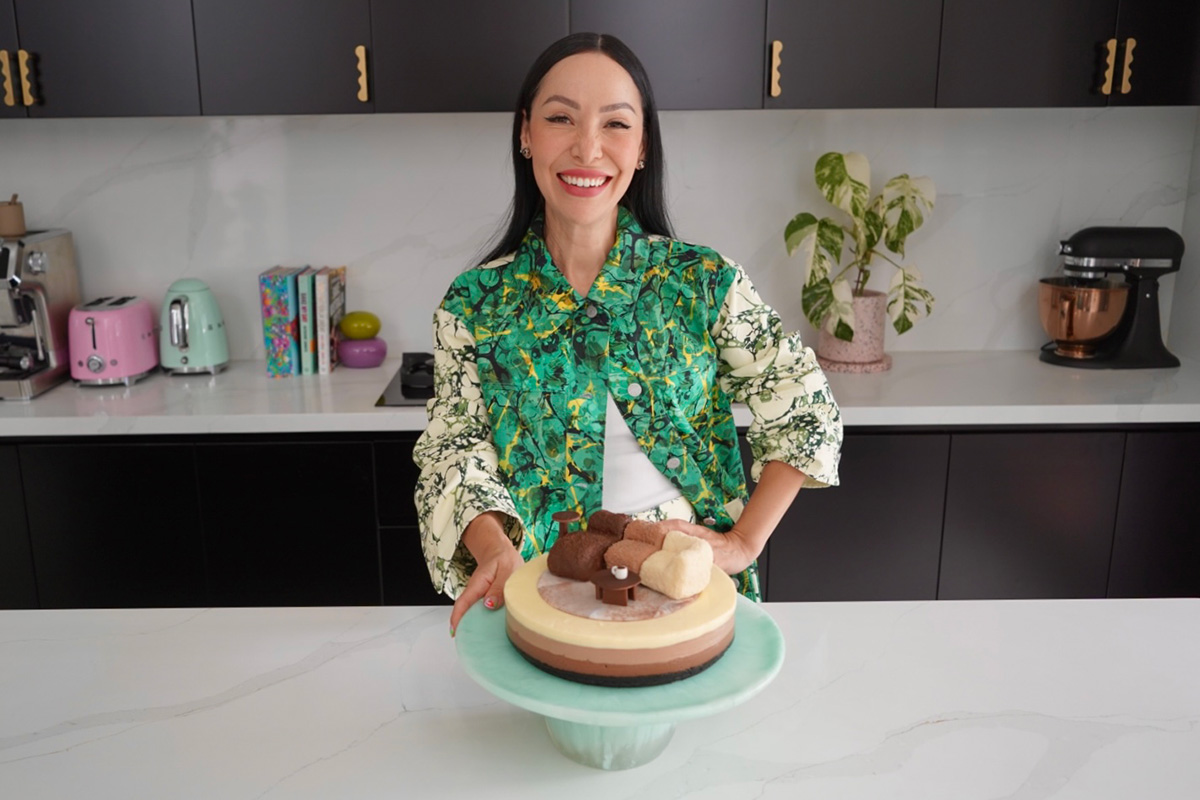 Cake artist Katherine Sabbath stands in a contemporary kitchen wearing a green patterned shirt, smiling with her layered dessert inspired by the modular King Living 1977 Sofa, echoing its curves and tones.