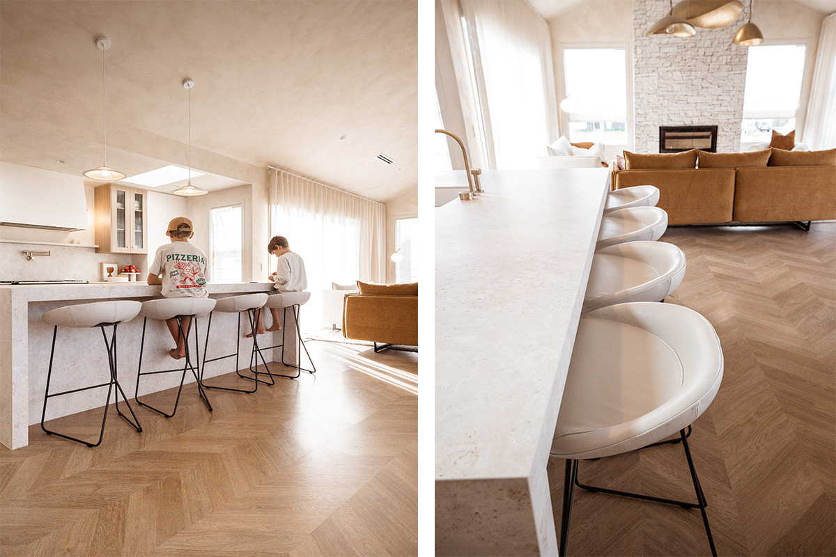 Two children seated at a neutral, marble kitchen island on white leather counter stools, with a view towards a tobacco-toned sofa in the living area.